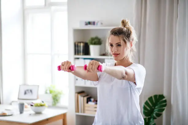 A young woman with dumbbells doing exercise in bedroom indoors at home.