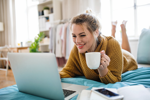 Happy young woman with laptop and coffee lying on bed indoors at home, relaxing.