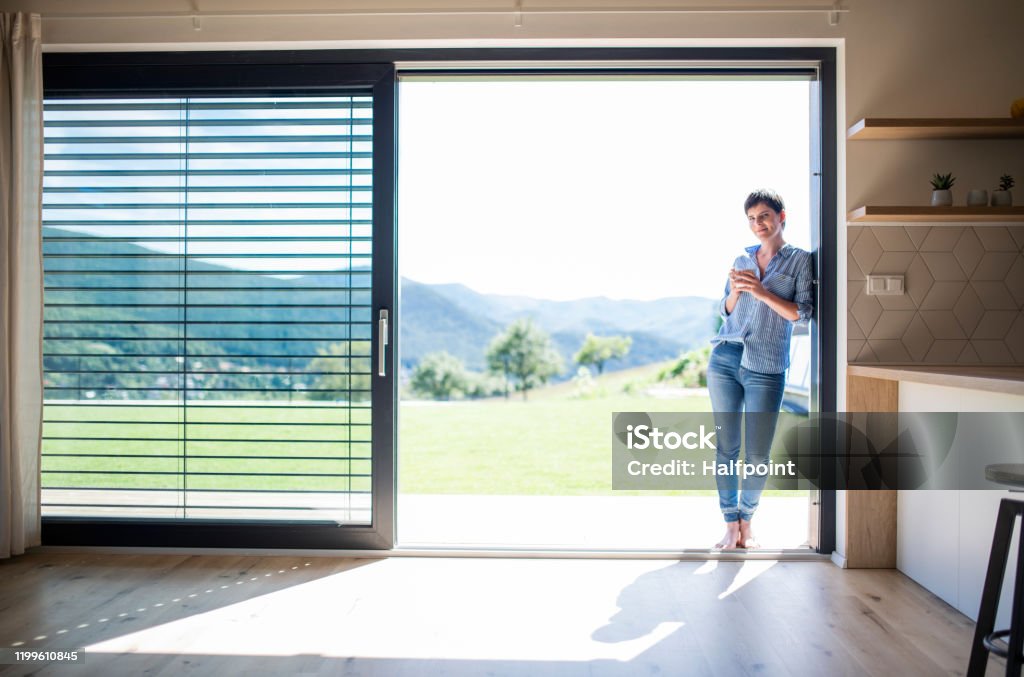 Front view of young woman with coffee standing by patio door at home. Front view of young woman with coffee standing by patio door at home. Copy space. Door Stock Photo