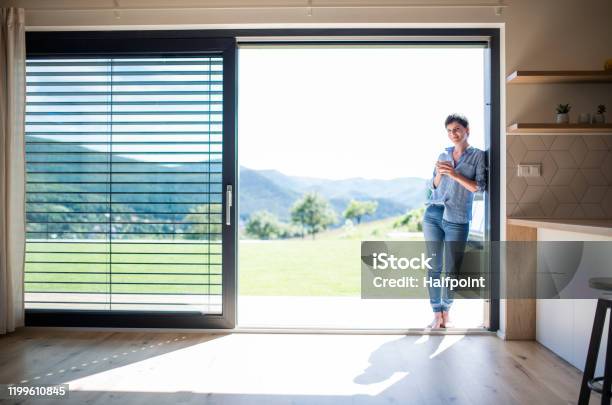 Front View Of Young Woman With Coffee Standing By Patio Door At Home Stock Photo - Download Image Now