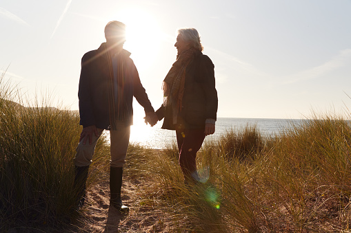 Silhouette Of Loving Senior Couple Holding Hands As They Walk Along Coast Path Against Flaring Sun