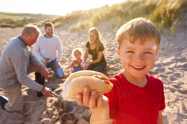 Grandfather Cooking As Multi-Generation Family Having Evening Barbecue Around Fire On Beach Vacation Grandfather Cooking As Multi-Generation Family Having Evening Barbecue Around Fire On Beach Vacation family bbq beach stock pictures, royalty-free photos & images