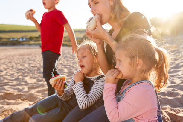 Mother With Children Eating Hot Dogs Sitting On Sand At Beach Barbecue Mother With Children Eating Hot Dogs Sitting On Sand At Beach Barbecue family bbq beach stock pictures, royalty-free photos & images