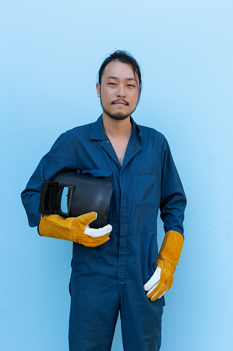 Retrato de soldador asiático con guantes de cuero sosteniendo casco de soldadura aislado en fondo azul photo