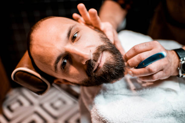 istock Young bearded man getting his beard retouched with a razor blade in a barber shop 1199622387