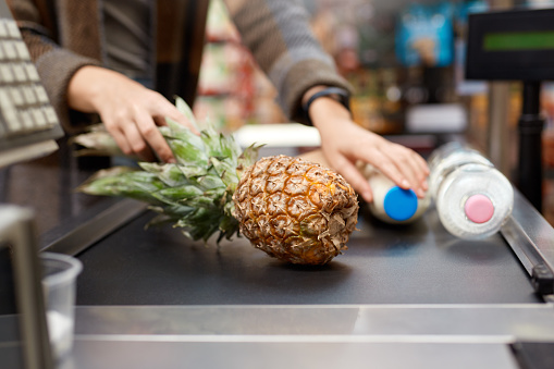 Cropped view of young adult woman shopping in supermarket, putting pineapple, bottle with milk and water on checking desk