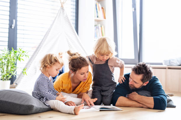 familia joven con dos niños pequeños en el interior del dormitorio leyendo un libro. - little girls small blond hair child fotografías e imágenes de stock