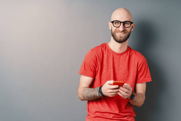 Handsome hairless Caucasian smiling man with beard, glasses, red T-shirt holding phone in hands on gray studio background stock photo