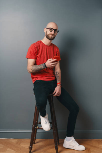 istock Handsome hairless man with beard,texting while holding smartphone in his tattooed arm on gray studio background 1199628439
