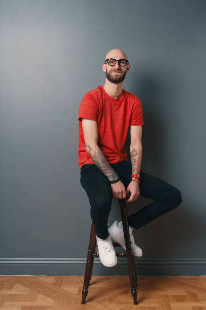 istock Young smiling man with beard and glasses sitting on a tall chair on gray studio background 1199628448