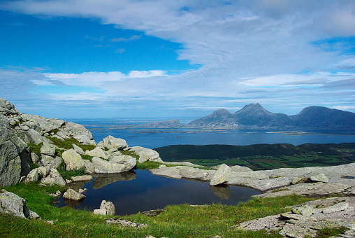 View to small lake by hiking trail in the mountain range of Seven sisters on sunny summer day against the background of Donna island surrounded by island group in blue Norwegian sea. Green valley of Alsta island seen below