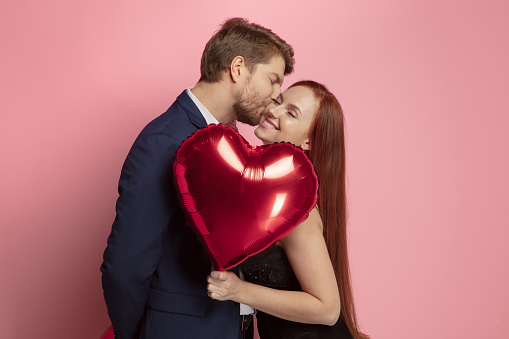 Happy holding balloons shaped hearts. Valentine's day celebration, happy caucasian couple on coral background. Concept of human emotions, facial expression, love, relations, romantic holidays.