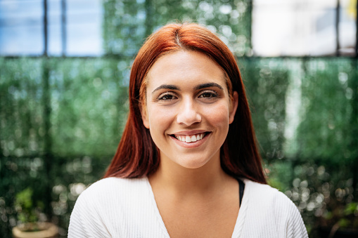 Close-up of Hispanic woman in early 20s with long red hair and casual white top smiling at camera in open-air cafe.