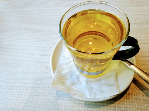 Cup of tea, glass cup, saucer and spoon, light wood background. Close-up high angle view, copy space on the left.