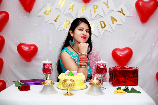 Young beautiful birthday girl of Indian ethnicity sitting with birthday cake with decorative background.