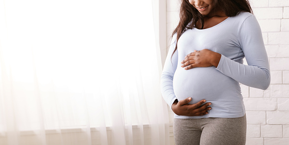 Black woman enjoying her pregnancy, hugging her tummy next to window at home, cropped, panorama with copy space