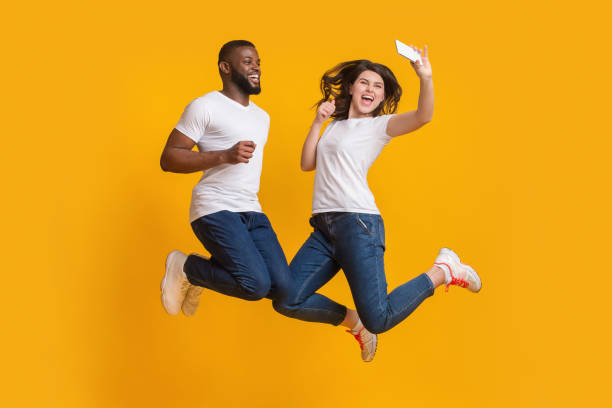 楽しい異人種間のカップルは、空気中でジャンプし、スマートフォンで自分撮りを取ります - couple indoors studio shot horizontal ストックフォトと画像