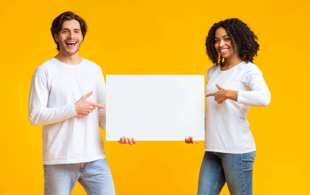istock Joyful interracial couple pointing at white blank placard in their hands 1199670152