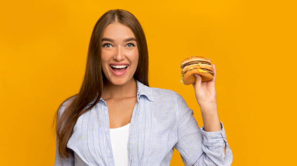 istock Cheerful Lady Holding Burger Standing On Yellow Studio Background, Panorama 1199670577