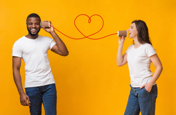 Romantic woman making love declaration to her boyfriend with tin phone Be My Valentine. Loving young woman sending romantic message to her afro boyfriend with tin phone, happy couple posing together over yellow background string telephone stock pictures, royalty-free photos & images