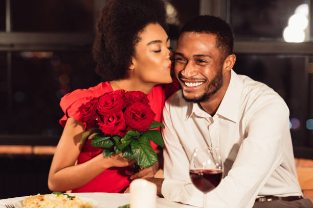 fidanzata che bacia il fidanzato dopo aver ricevuto rose durante l'appuntamento al ristorante - men african descent giving flower foto e immagini stock