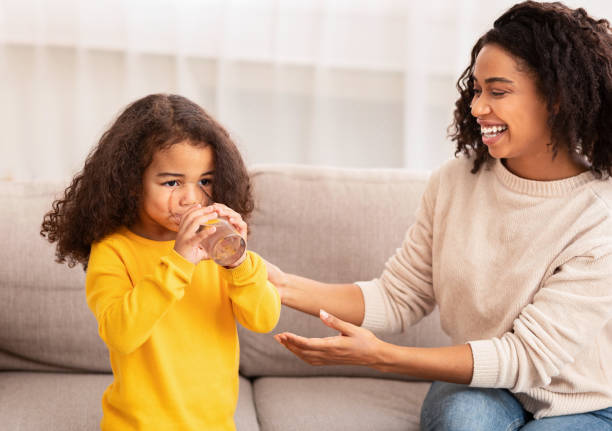 woman giving daughter glass of water sitting on sofa indoor - glasses child cute offspring imagens e fotografias de stock