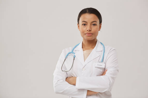 Young Female Doctor Posing on White Waist up portrait of beautiful African-American nurse posing confidently while standing with arms crossed against white background, copy space lab coat photos stock pictures, royalty-free photos & images