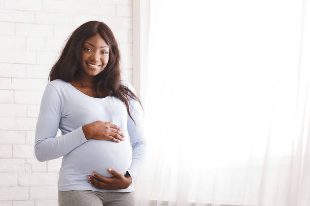 femme d'afro enceinte de sourire posant à la maison - abdomen adult affectionate baby photos et images de collection