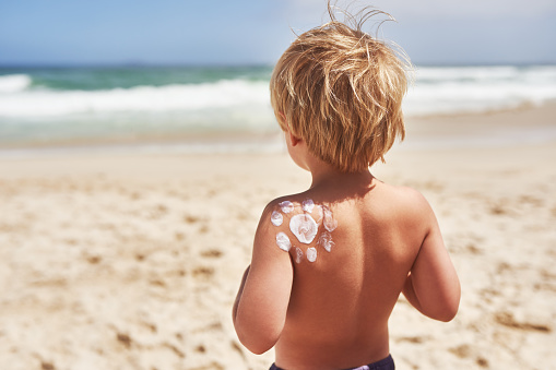 Rearview shot of a little boy standing at the beach with sunscreen lotion on his back