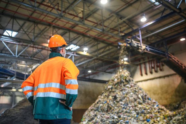 Worker Observing Processing of Waste at Recycling Facility Low angle rear view of young male worker in helmet, pollution mask, and reflective clothing observing waste falling from conveyor belt onto pile at facility. social responsibility photos stock pictures, royalty-free photos & images