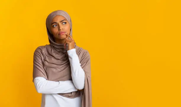 Portrait of doubtful afro muslim woman in headscarf touching chin, overthinking options and looking away over yellow background