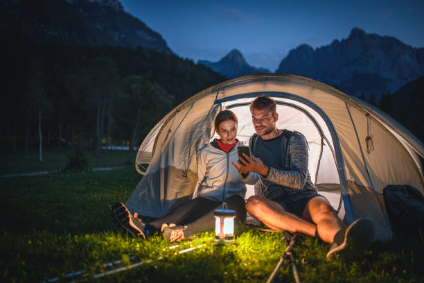 Close-up of young camping couple sitting on tent porch after day of hiking and checking smart phone with Julian Alps in background.