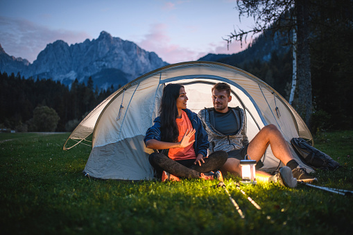Relaxed camping couple in 20s and 30s sitting on tent porch and talking at dusk with Julian Alps in background.