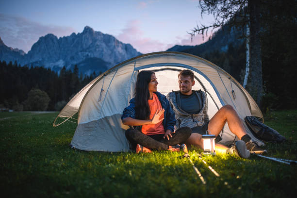 Young Couple Camping in Northwestern Slovenia Relaxed camping couple in 20s and 30s sitting on tent porch and talking at dusk with Julian Alps in background. lanterns at night stock pictures, royalty-free photos & images