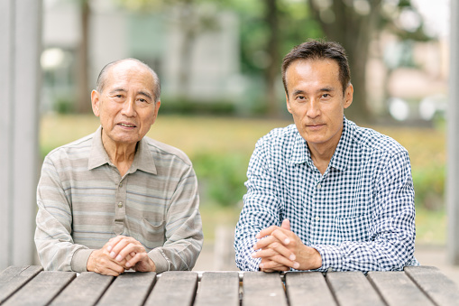 A portrait of a senior father with his mature adult son while sitting on a bench in a public park.