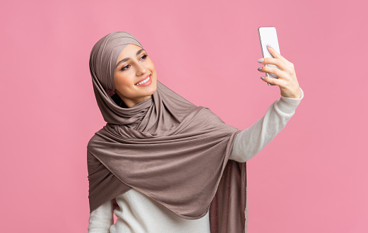 International selfie. Beautiful muslim girl wearing hijab taking self-portrait on smartphone over pink background.