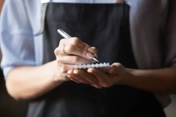 Waitress small cafe business owner wear apron uniform holding notepad pen write notes take order in restaurant, retail serving waiting staff, hospitality, good customer service concept, close up view