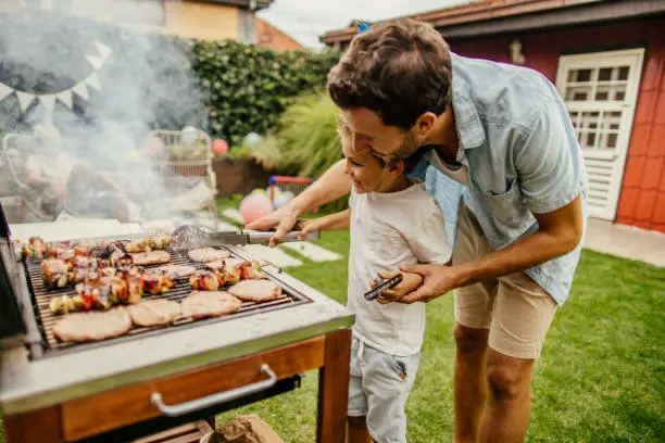 Photo of father and son grilling meat during the barbecue party in their yard
