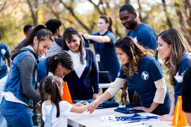 Mid adult volunteer shakes hands with girl at registration table While other people register, a mid adult volunteer reaches across the table to shake hands with a young girl.  Other volunteers smile and watch. social responsibility photos stock pictures, royalty-free photos & images