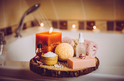 Home spa products on wooden tray: bar of soap, bath bomb, aroma bath salt, essential and massage oils, candle burning, rolled towel inside bathroom by tub running water. Modern Instagram style filter.