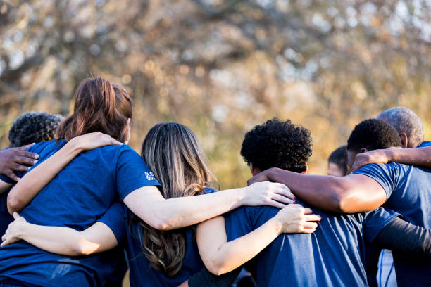 Friends linking arms in unity Diverse group of friends cleanup a park during a charity event. They are standing with their arms around one another. social services stock pictures, royalty-free photos & images