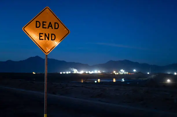 Dark night sky with a reflective sign at a dead end road. In the background is a a view of a construction dig site with a low depth of field