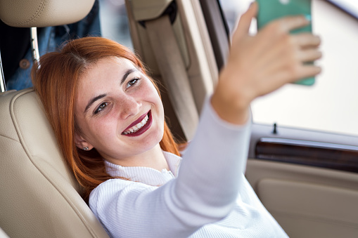 Young redhead woman driver taking selfies with her mobile phone sitting behind the wheel of the car in rush hour traffic jam.
