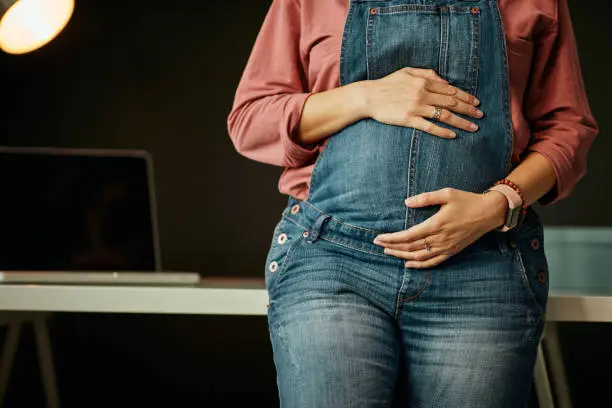 Close up of caucasian pregnant woman standing in home office, leaning on desk and touching belly.