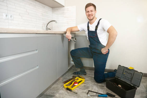 young smiling man plumber in uniform fixing the sink with adjustable spanner in his hand working in the kitchen professional plumbing repair service stock photo