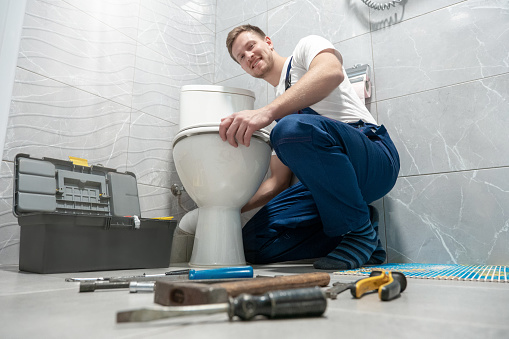 smiling man plumber in uniform repairing toilet bowl using instrument kit looks happy professional repair service.