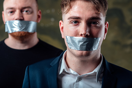 two young man with mouth and lips sealed covered with grey adhesive tape in censorship coerced freedom of speech and forced silence and secrecy concept isolated on dark  background, close up