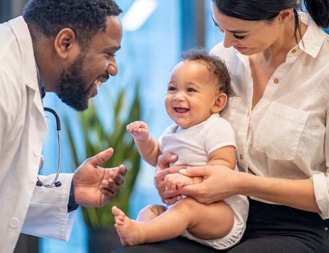 A Hispanic Mother brings in her mixed race son for a wellness check-up.  She has the baby sitting on her lap with her hands firmly around his waist.  The African male doctor  is reaching out to hold the baby's hand.  The doctor is laughing and engaging the young boy.