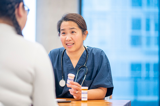An Asian nurse meets with a young Native girl to talk about her new medication.  The girl is dressed casually in a white shirt, has braided pigtails in her hair, and her back towards the camera.  The nurse is wearing blue scrubs with a stethoscope around her neck and is talking to the girl about the possible side effects of the new medication.