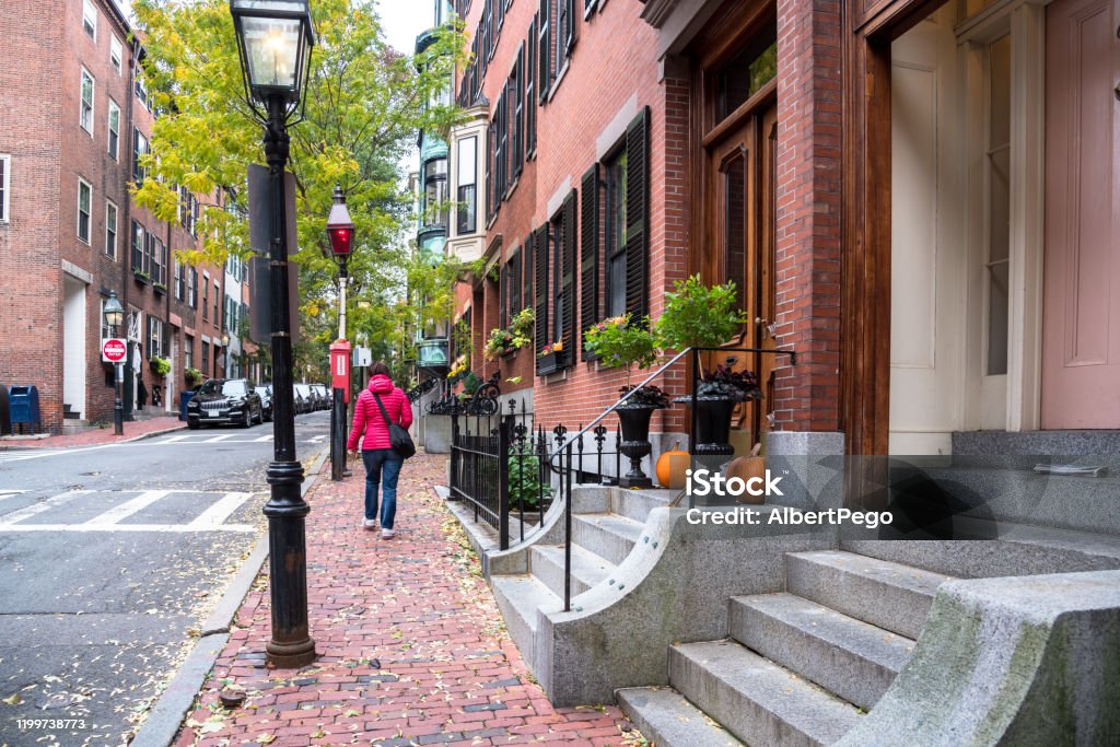 Woman walking on the sidewalk of a historic residential district Woman walking along a brick sidewalk lined with trees and traditional American row houses on a cloudy autumn day. Beacon hill, Boston, MA, USA. Walking Stock Photo